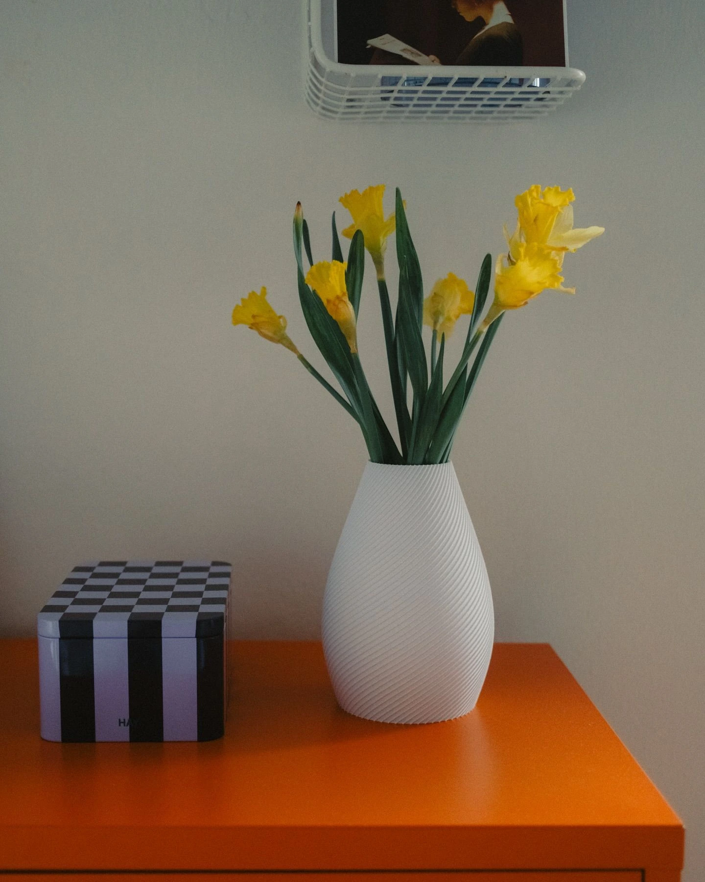 White ribbed vase with yellow daffodils on an orange sideboard next to a checkerboard box